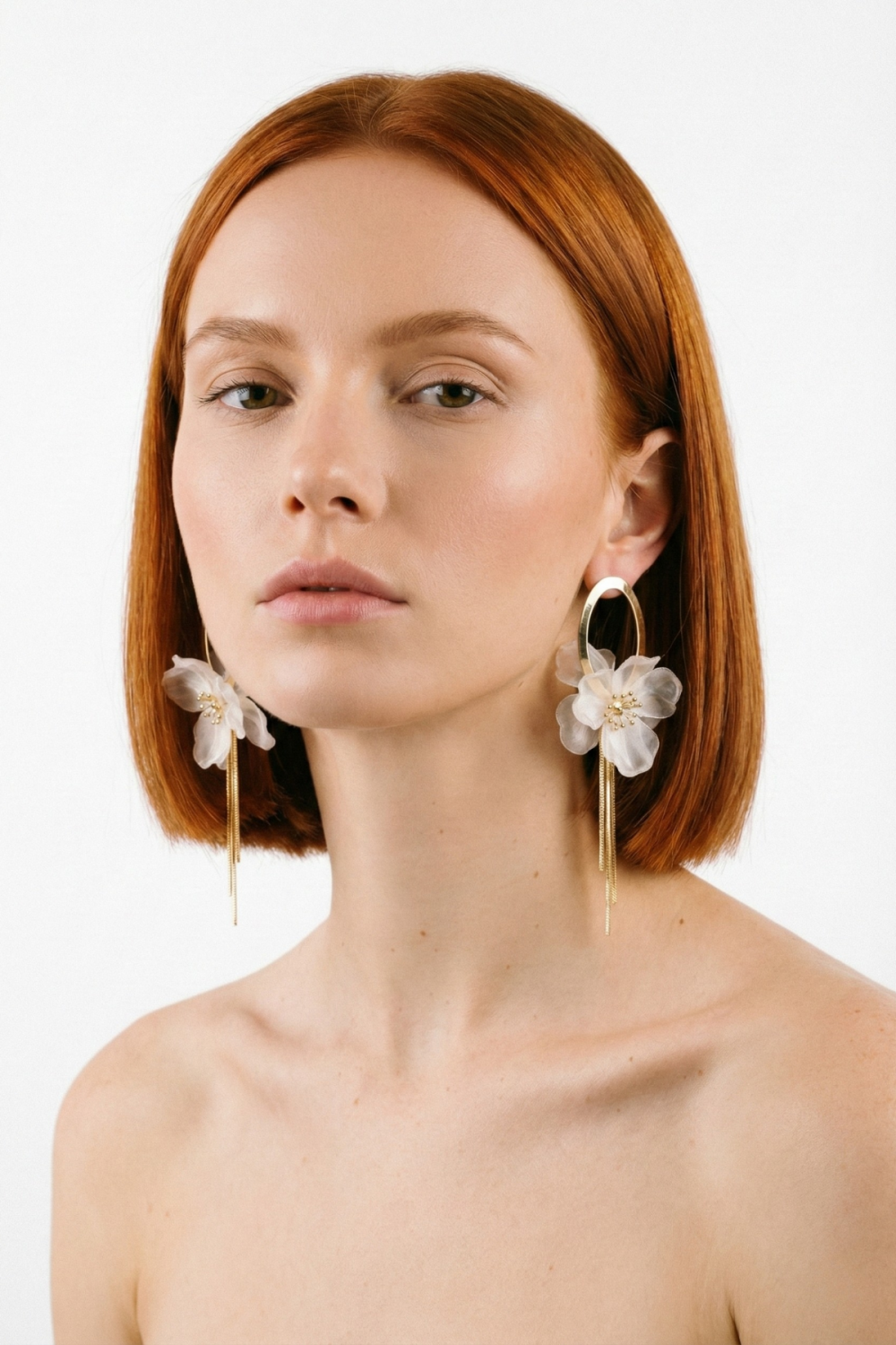 Boucles d’oreilles Louvre en longue suspension de 11 cm, composées d’un ovale doré et d’une fleur blanc cassé en matière nacrée, portées par une femme rousse au carré sur fond blanc. Bijou architectural et raffiné.