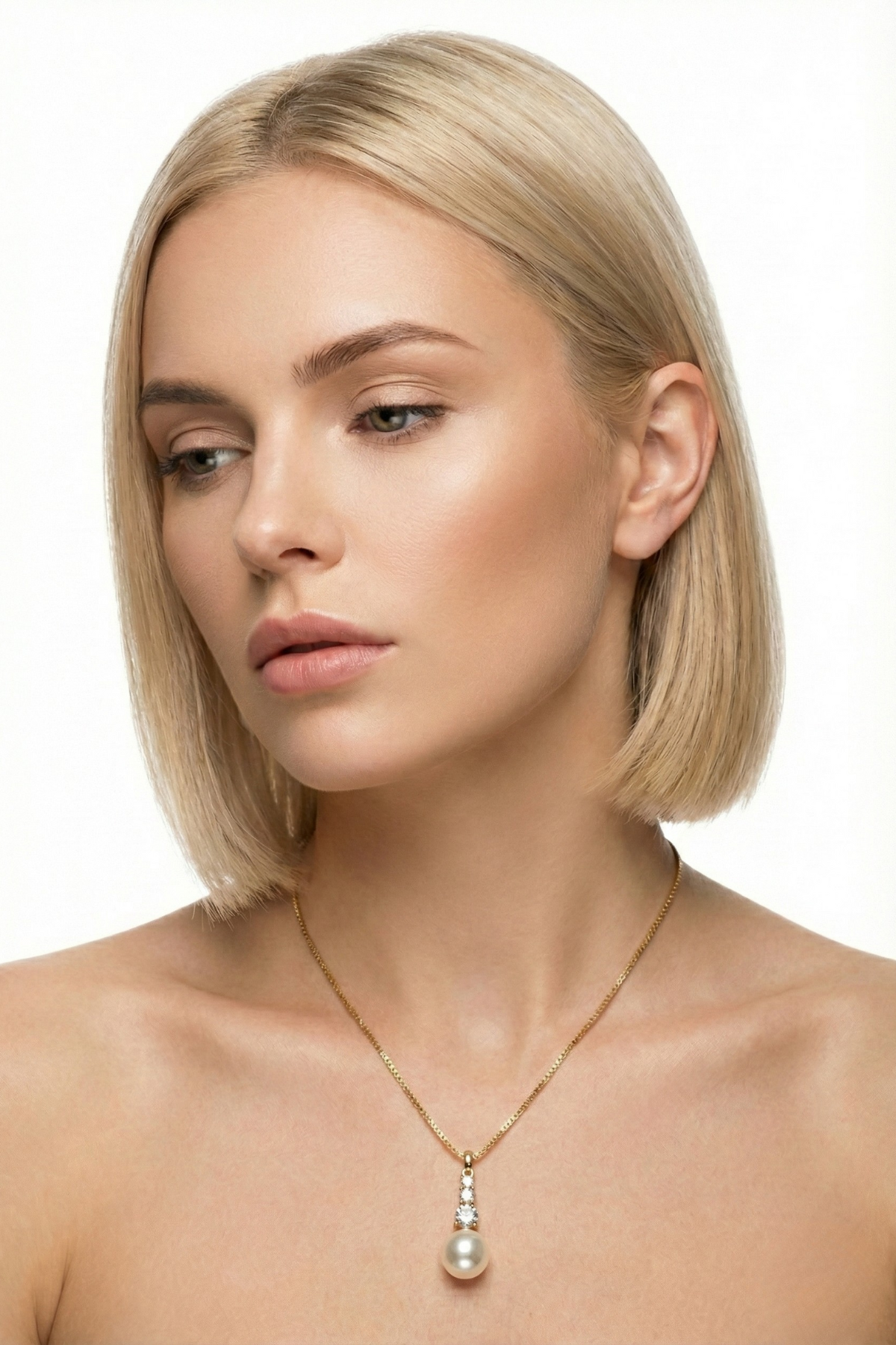 Collier Lutétia doré en acier inoxydable, composé d’une fine chaîne et d’un pendentif avec strass scintillants surmontant une perle fantaisie nacrée, porté par une femme blonde au carré sur fond blanc