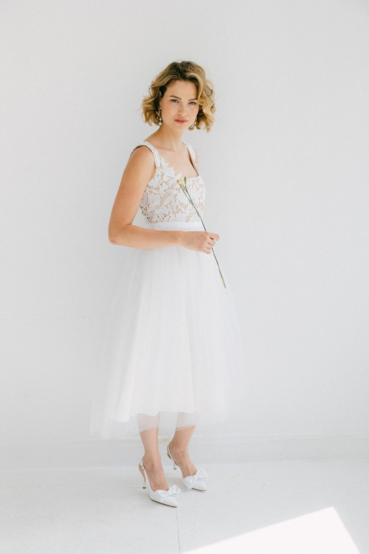 Mariée portant la robe Lana mi-longue en guipure blanche et tulle aérien, silhouette légère et élégante dans un studio lumineux.