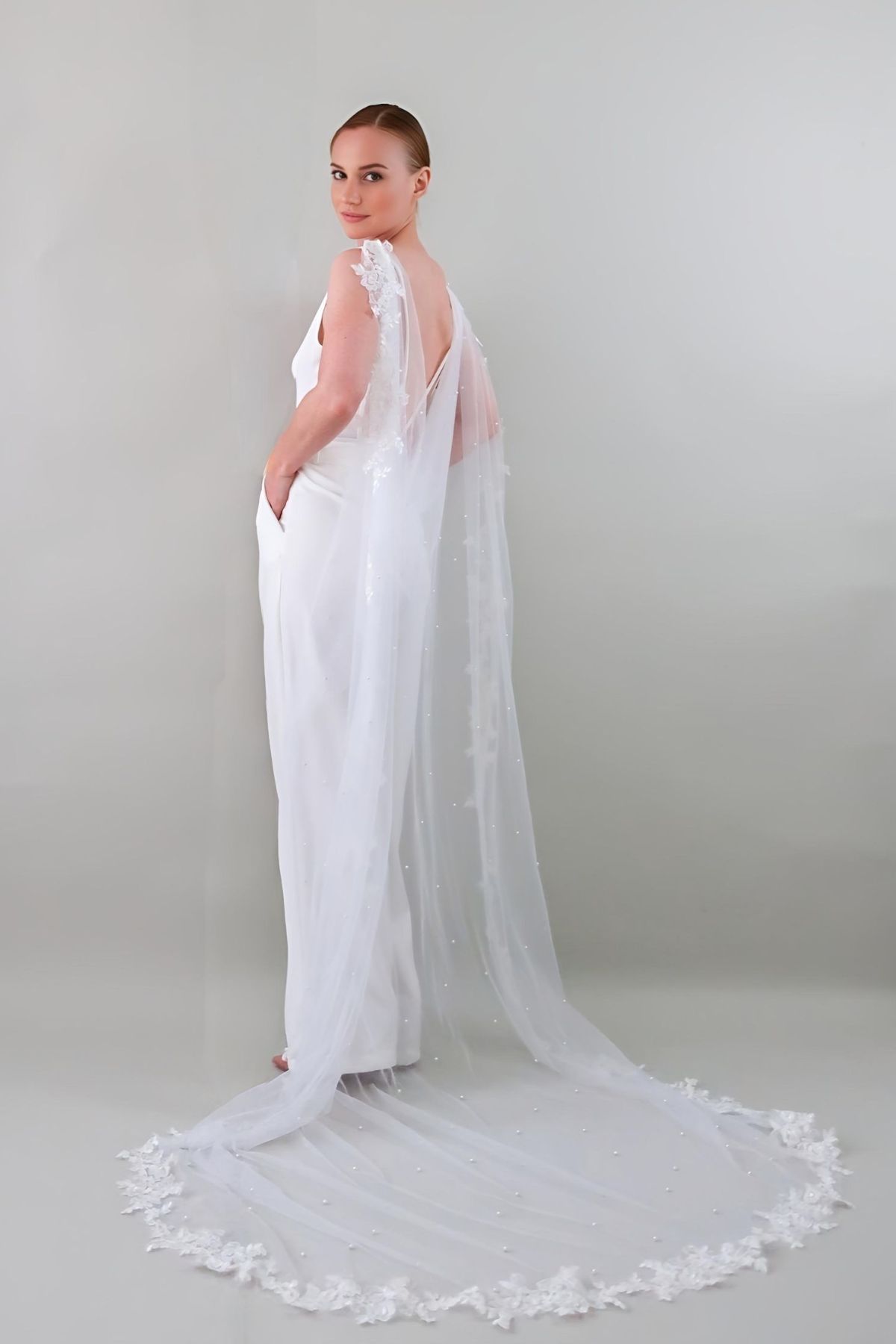 Mariée portant la cape Capri en tulle fluide bordée de dentelle et perles, accessoire amovible élégant pour robe de mariée.