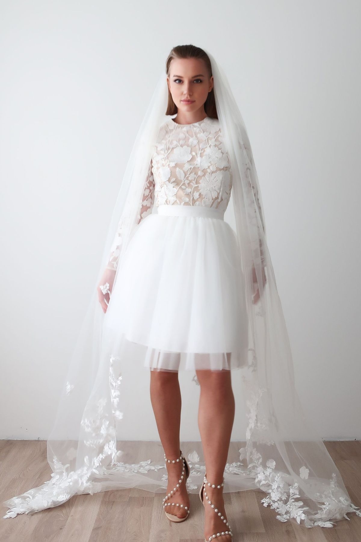 Mariée portant le voile cathédrale Victoria en tulle fluide et dentelle florale, posé sur une robe courte en dentelle, ambiance élégante et lumineuse.